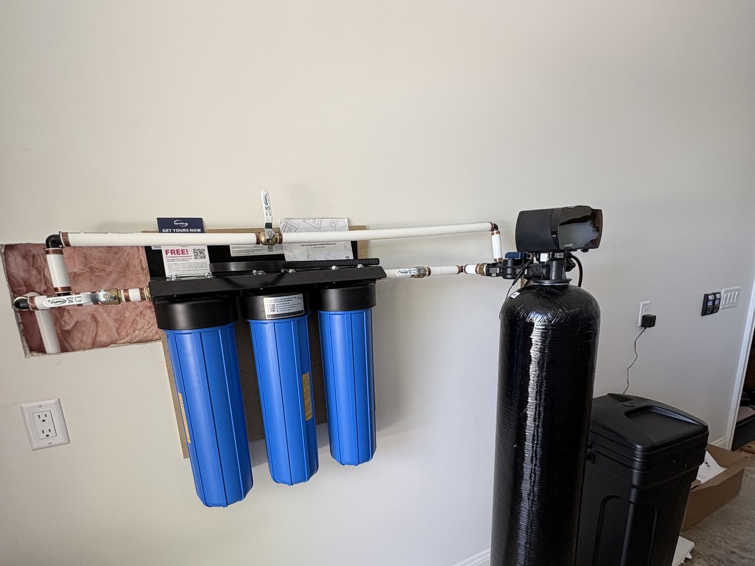 Three-stage whole-house water filtration system with cartridge housings and a black water softener tank installed on a wall